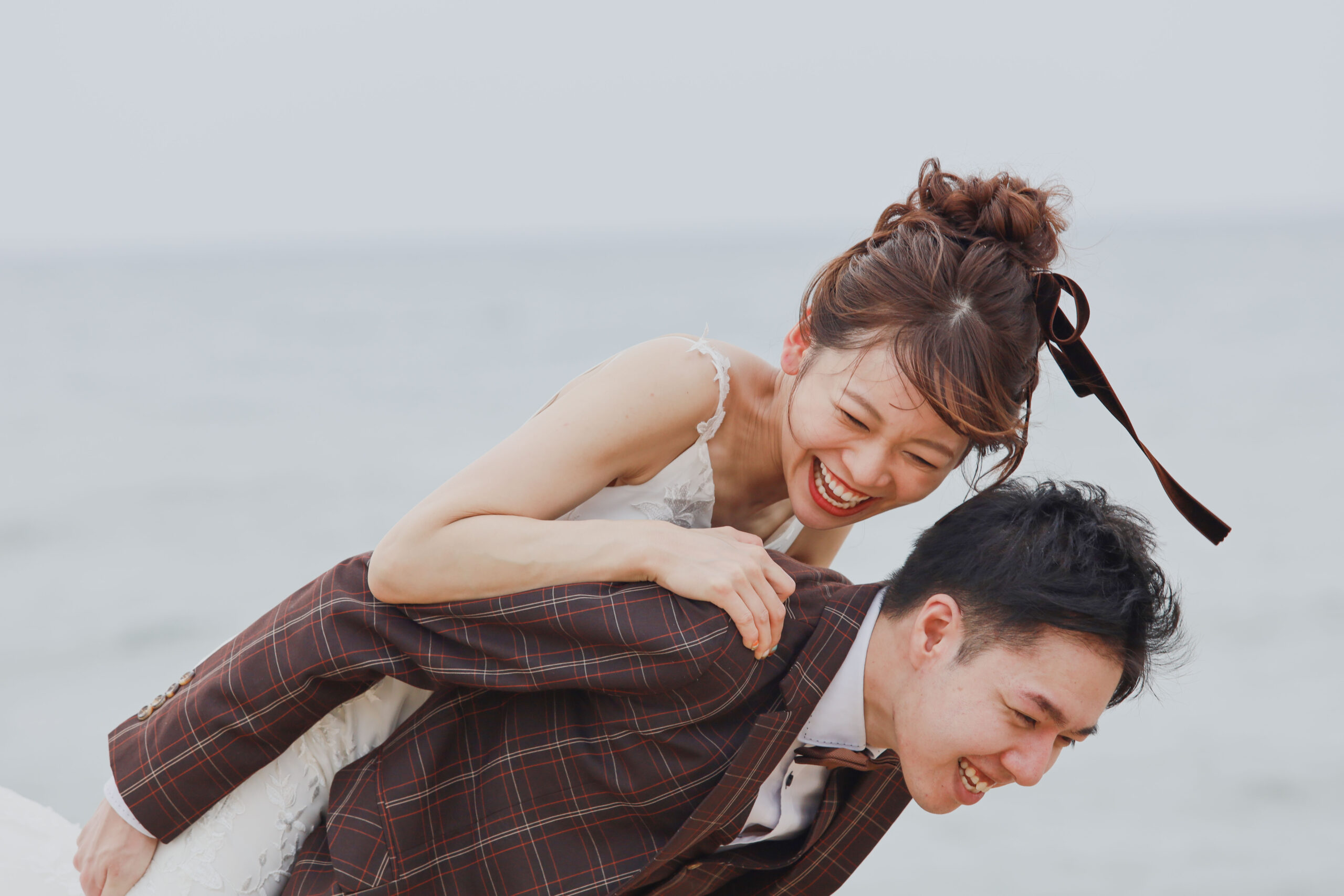 富山県射水市にあるウエディングフォトスタジオシャトンで行った新郎新婦が海辺でおんぶをして笑い合っているフォトウェディング